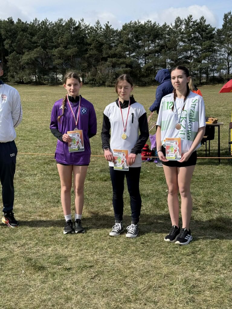 Trzy dziewczyny z medalami i voucherami stoją na trawie na tle lasu, prawdopodobnie podczas wręczenia nagród sportowych.