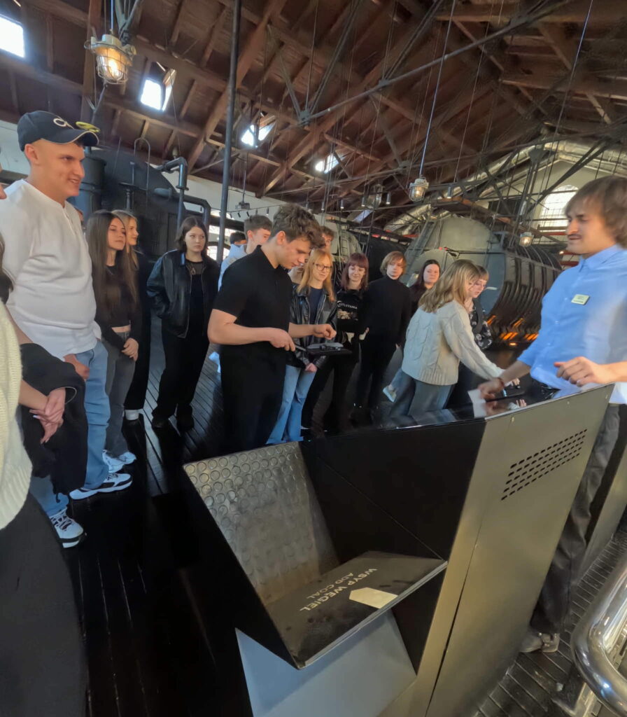 Grupa młodych ludzi uczestniczy w oprowadzaniu w industrialnym wnętrzu, skupiając się na makiecie lub urządzeniu opatrzonym napisem „WSBIV WEIGEL POD COAL”.