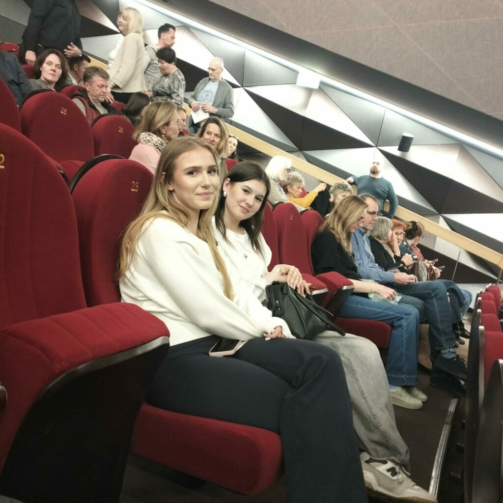 Grupa osób siedzi na czerwonych fotelach w kinie lub teatrze, dwie młode kobiety na pierwszym planie uśmiechają się do aparatu.