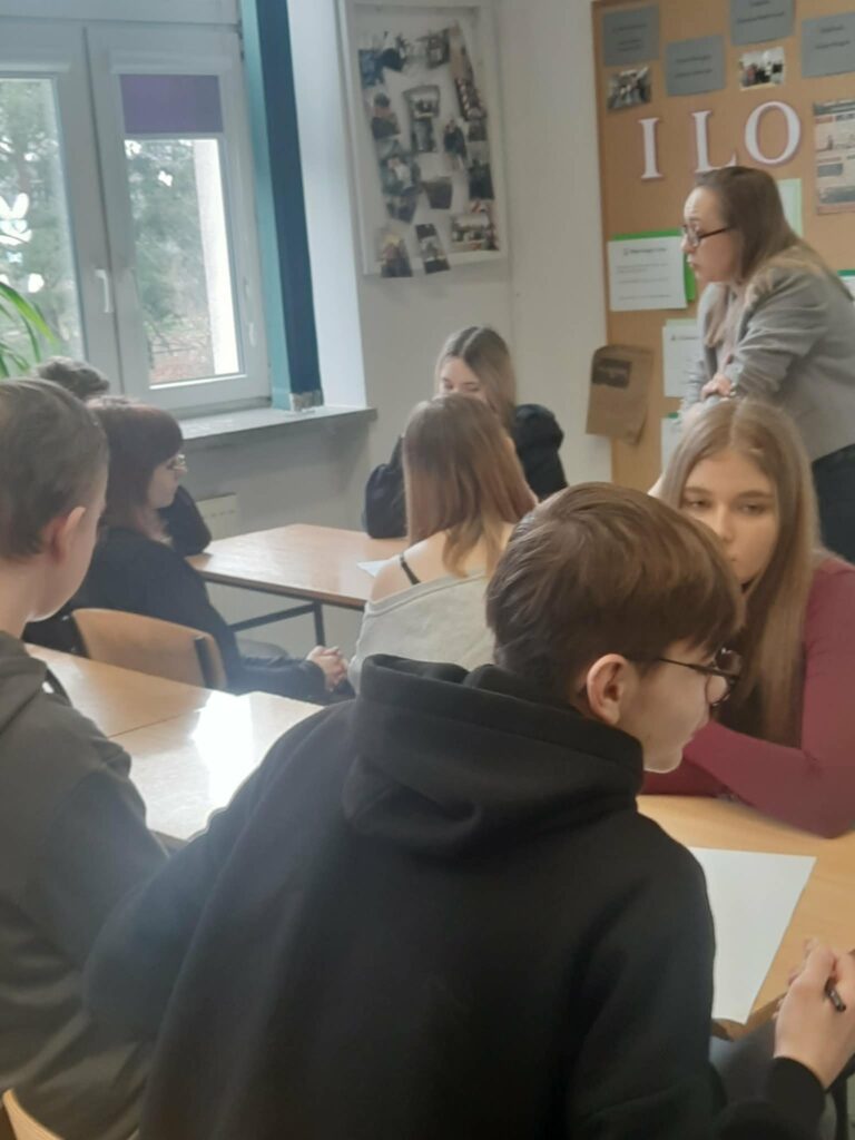 Grupa młodzieży siedzi przy stołach w klasie, prowadząc dyskusję pod opieką nauczycielki.