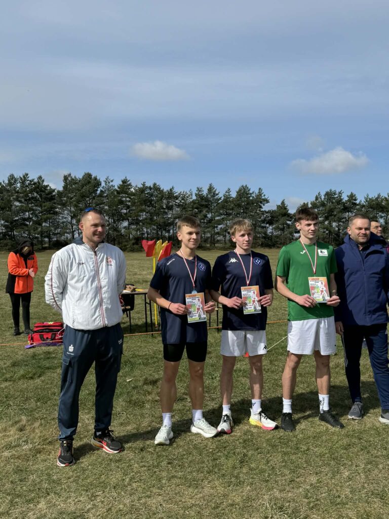 Trzech młodych sportowców z medalami i dyplomami stoi na tle boiska i drzew, towarzyszą im dwaj dorośli mężczyźni.