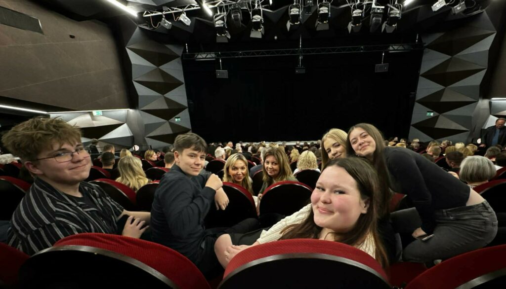 Grupa młodych osób siedzi na czerwonych fotelach w sali kinowej lub teatralnej, uśmiechając się do aparatu.