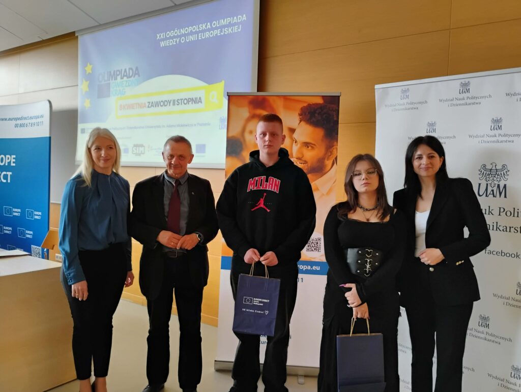 Pięć osób pozujących do zdjęcia podczas XXI Ogólnopolskiej Olimpiady Wiedzy o Unii Europejskiej, z dwoma młodymi uczestnikami trzymającymi torby z nagrodami.