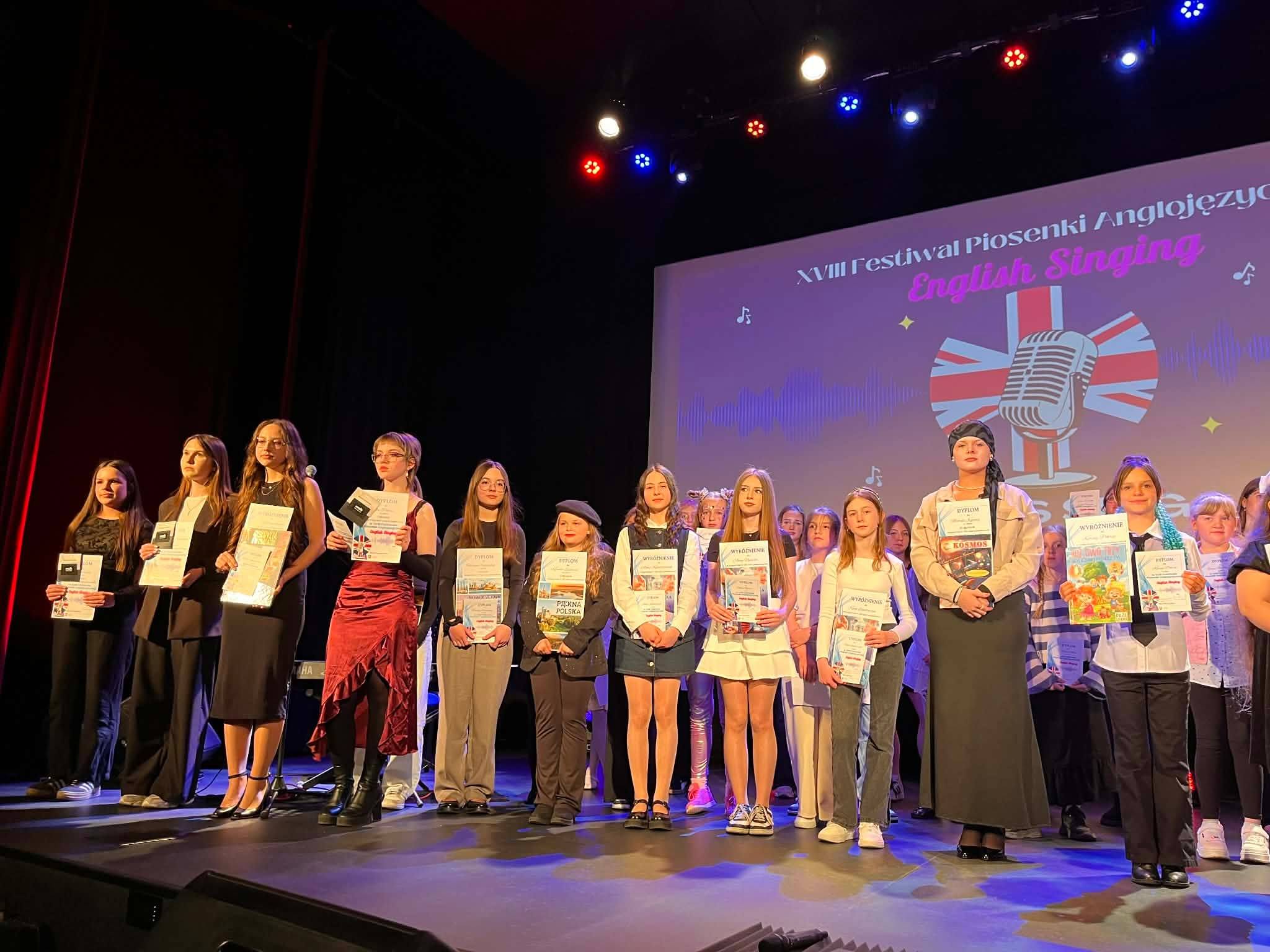 Grupa młodych uczestników stoi na scenie z dyplomami i nagrodami podczas XVIII Festiwalu Piosenki Anglojęzycznej English Singing.