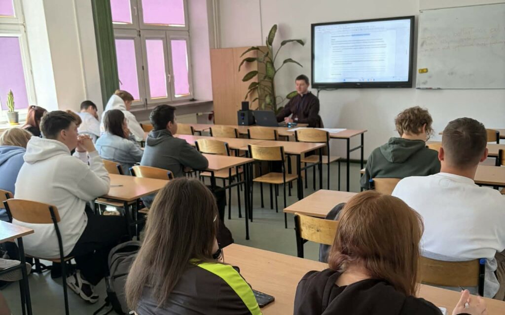 Uczniowie siedzą w ławkach i słuchają nauczyciela prowadzącego lekcję przy tablicy interaktywnej w klasie.