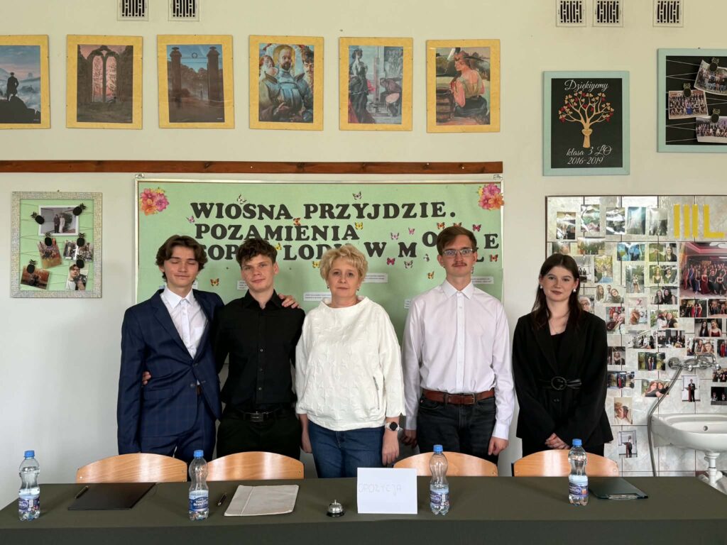 Grupa czterech młodych osób i kobieta stoją przed tablicą z napisem „Wiosna przyjdzie. Pozamienia poglądy w moście”.