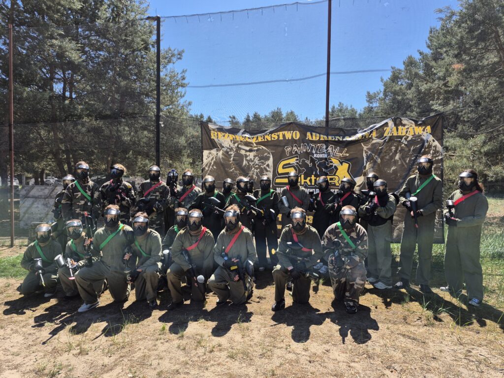 Grupa osób w strojach i maskach ochronnych do paintballa, trzymająca markery, stoi na polu przed banerem z napisem dotyczącym paintballa.