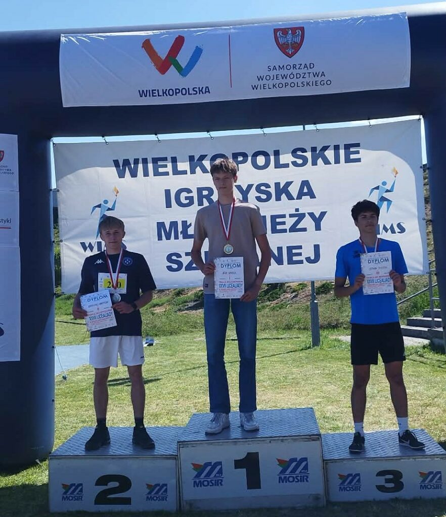 Trzech chłopców stoi na podium z medalami i dyplomami, zwycięzcy wielkopolskich igrzysk młodzieży szkolnej.
