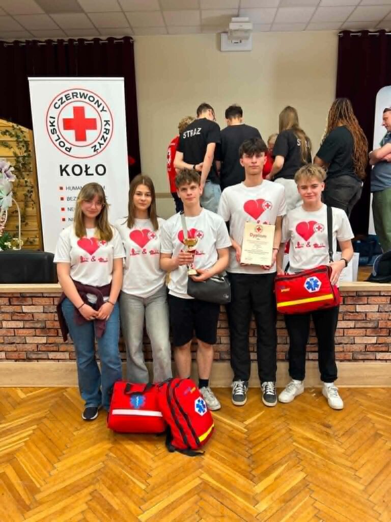 Grupa uczestników i organizatorów XXXII Ogólnopolskich Mistrzostw Pierwszej Pomocy szkolnego etapu powiatowego z plecakami i dyplomem na tle banerów Polskiego Czerwonego Krzyża.