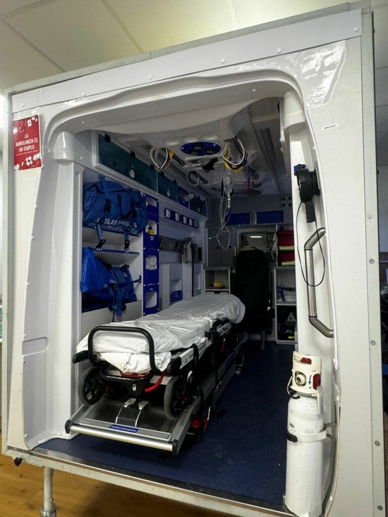 Wnętrze ambulansu z noszami, wyposażonego w niebieskie torby medyczne i sprzęt ratunkowy.