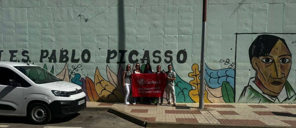 Grupa osób trzymająca czerwoną flagę z napisem "Carlos V" stoi na tle kolorowego muralu z napisem "Pablo Picasso" i portretem mężczyzny.