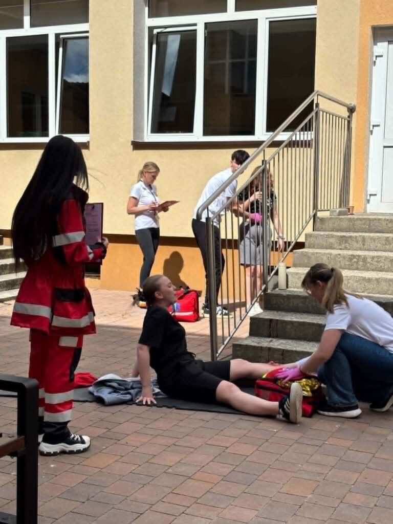 Grupa osób ćwiczy udzielanie pierwszej pomocy na zewnątrz budynku, jedna osoba symuluje poszkodowanego leżącego na ziemi, inni udzielają mu pomocy i notują obserwacje.
