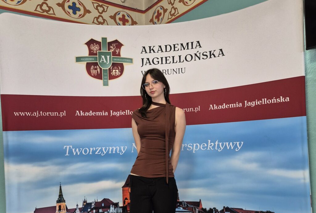 Młoda kobieta w brązowej bluzce i okularach stoi na tle banera Akademii Jagiellońskiej w Toruniu.