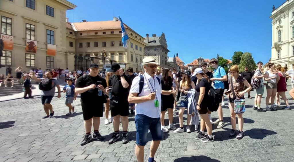 Grupa turystów z przewodnikiem trzymającym niebieską flagę stoi na placu otoczonym historycznymi budynkami w słoneczny dzień.