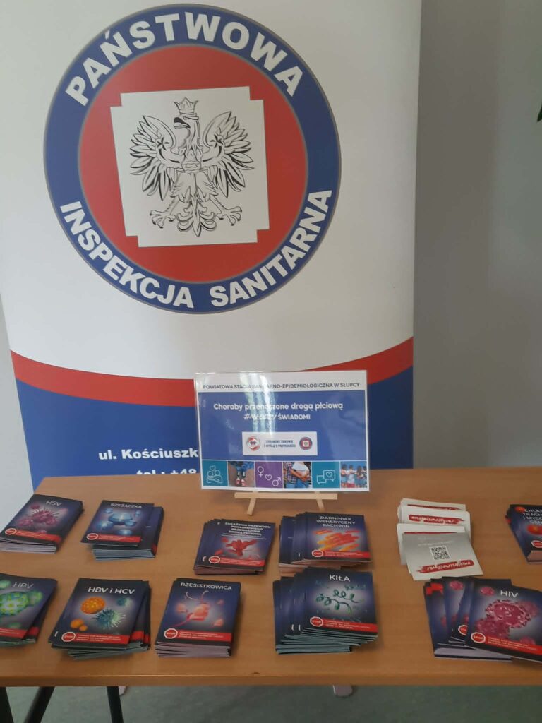 Materiały informacyjne o chorobach przenoszonych drogą płciową ułożone na stole, z logo Państwowej Inspekcji Sanitarnej w tle.