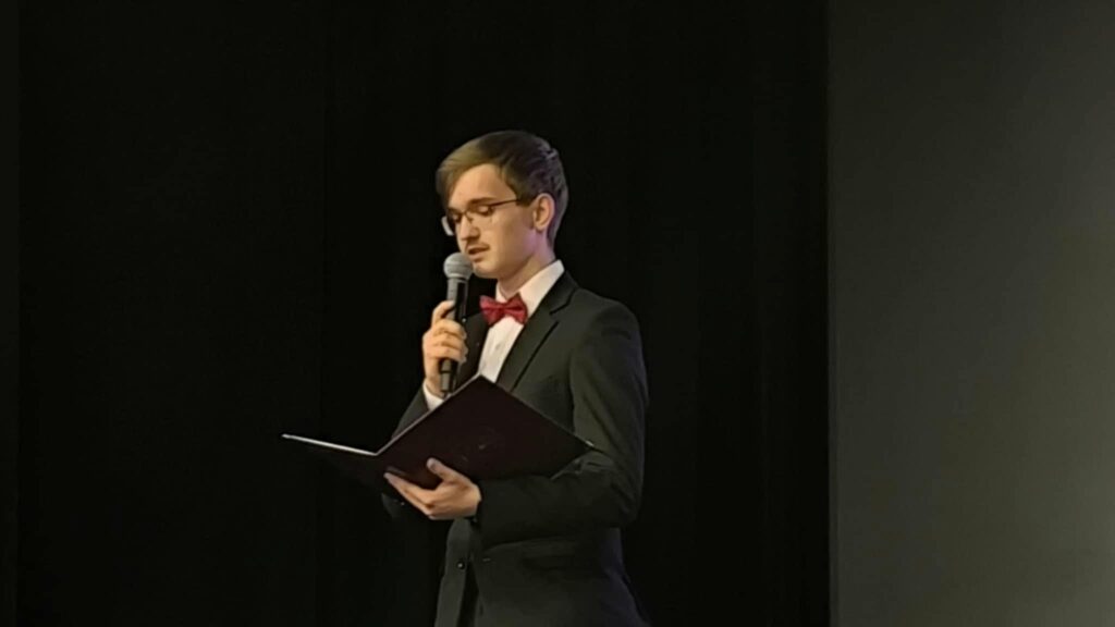 Młody mężczyzna w garniturze z muszką przemawia do mikrofonu, trzymając otwartą teczkę.