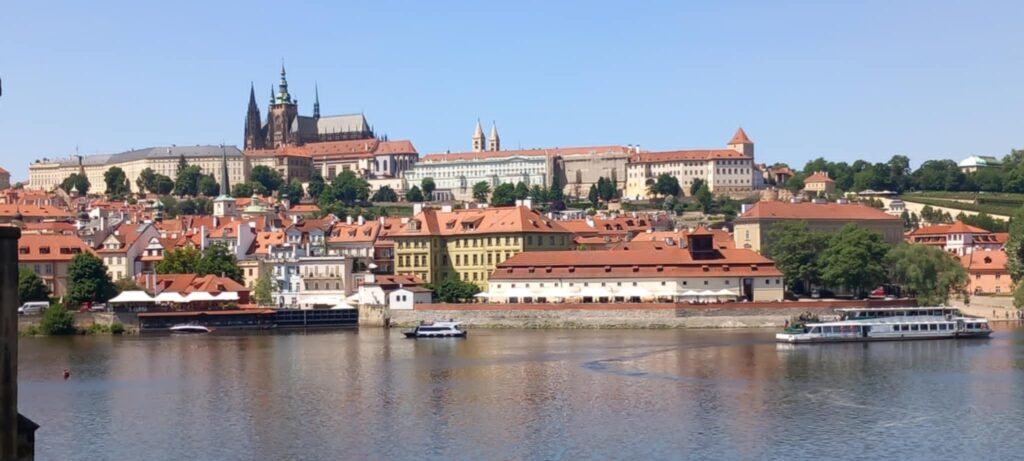 Panorama miasta z rzeką na pierwszym planie, liczne zabytkowe budynki i kościoły z czerwonymi dachami oraz statki wycieczkowe na wodzie.