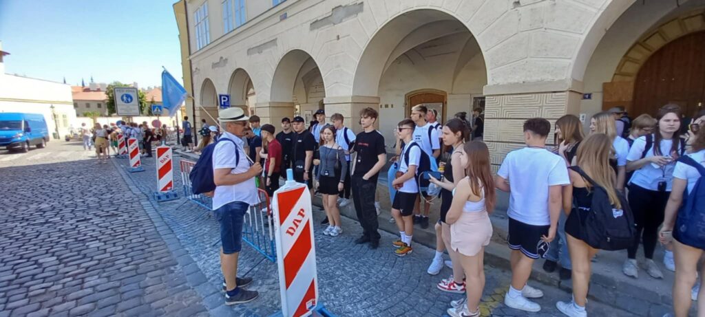 Grupa młodych ludzi stoi przy ogrodzeniu na brukowanym chodniku obok budynku z arkadami, a jeden mężczyzna z kapeluszem i plecakiem rozmawia z nimi.