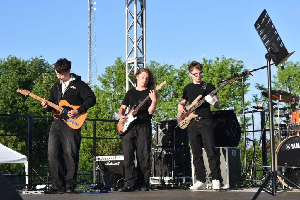 Trzech młodych muzyków gra na gitarach elektrycznych i basowej podczas koncertu na świeżym powietrzu przy słonecznej pogodzie.