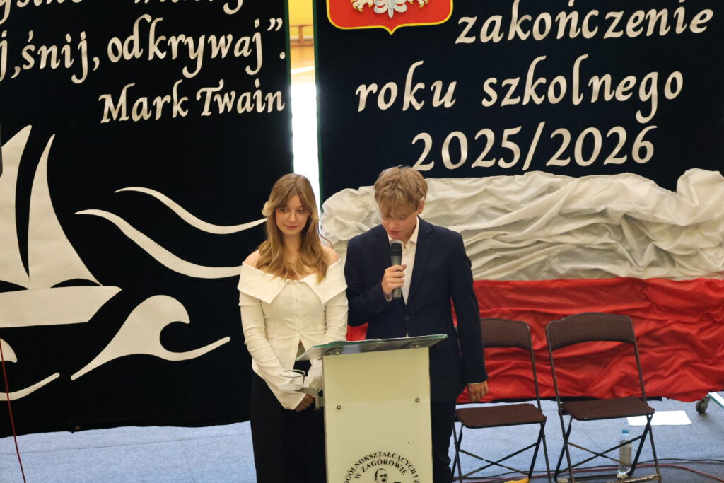 Dwóch młodych ludzi stoi przy mikrofonie na szkolnej scenie podczas zakończenia roku szkolnego 2025/2026, z tłem w narodowych barwach Polski i cytatem Marka Twaina.