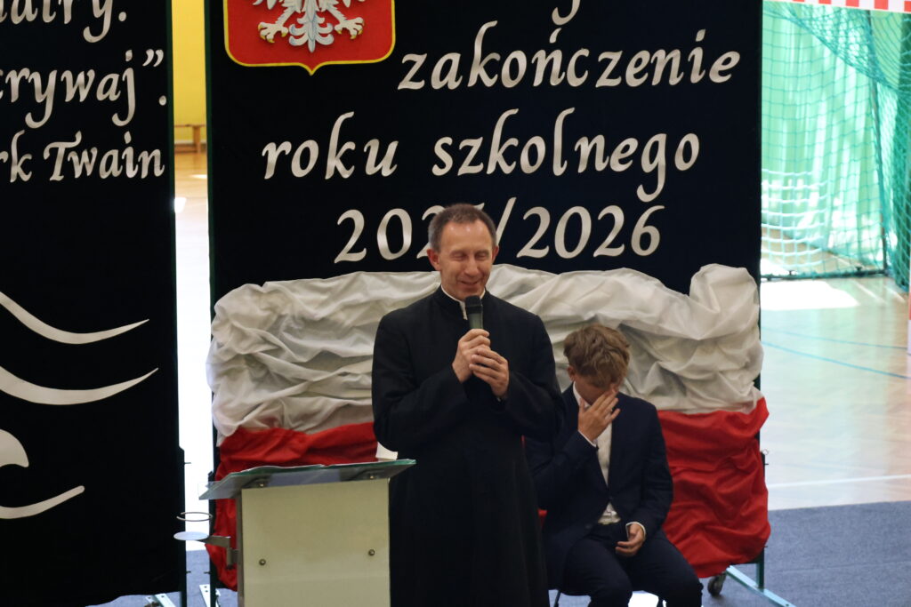 Ksiądz przemawia na zakończeniu roku szkolnego 2025/2026, obok siedzi chłopiec w garniturze zasłaniający twarz ręką.