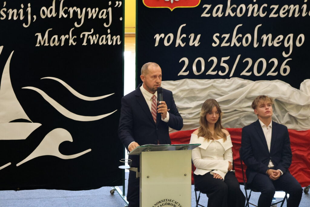 Mężczyzna w garniturze przemawia przy mównicy podczas uroczystości zakończenia roku szkolnego 2025/2026, obok siedzą dwie młode osoby.