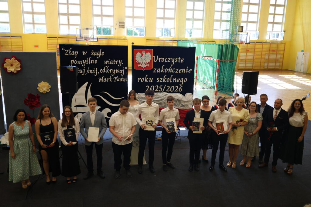 Uczniowie i nauczyciele stojący na scenie w hali sportowej podczas uroczystego zakończenia roku szkolnego 2025/2026.