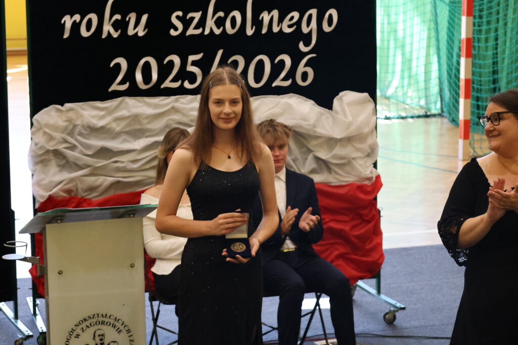 Młoda kobieta w czarnej sukni odbiera medal podczas uroczystości szkolnej na rok 2025/2026, a widoczni wokół ludzie klaszczą i uśmiechają się.