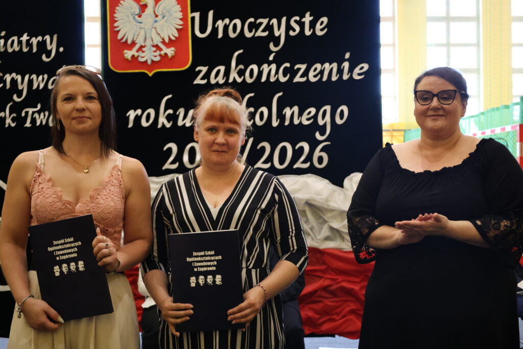 Troje kobiet stoi na tle baneru z napisem „Uroczyste zakończenie roku szkolnego 2025/2026”, dwie z nich trzymają czarne teczki z napisem „Zespół Szkół Ogólnokształcących i Zawodowych w Zagórowie”.