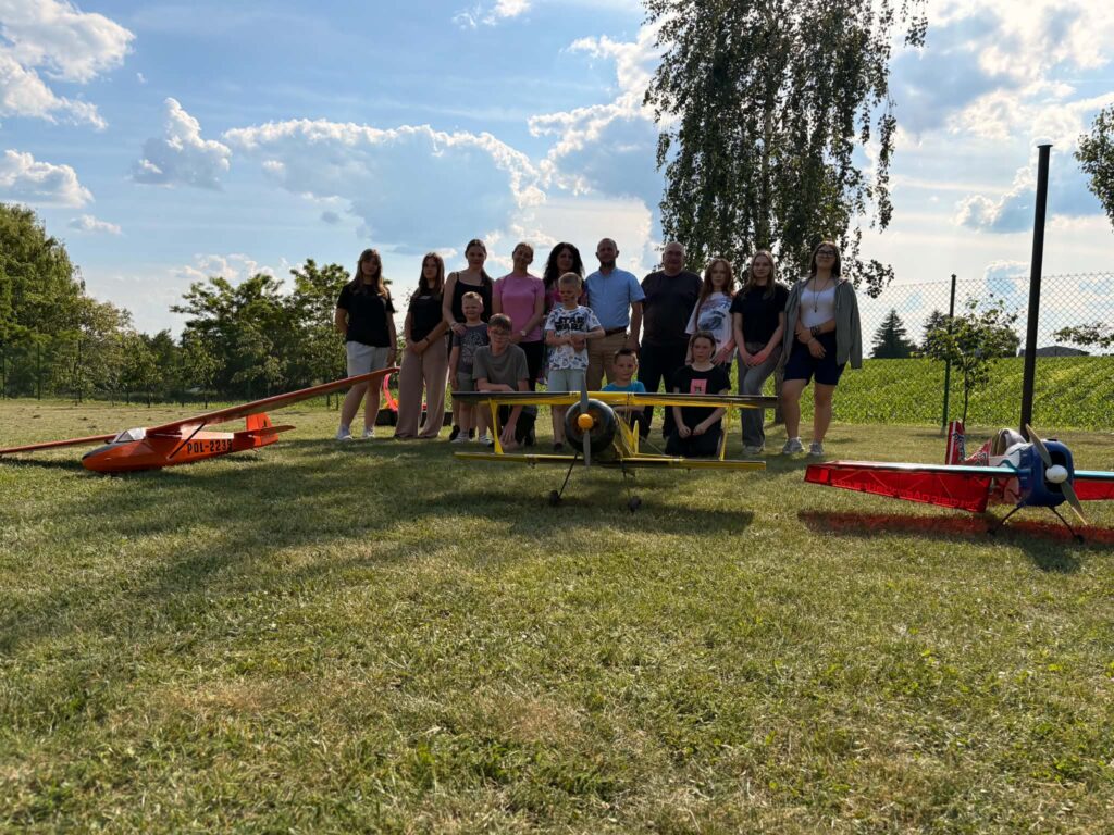 Grupa osób stoi na trawie za trzema modelami samolotów zdalnie sterowanych rozstawionymi na polu.