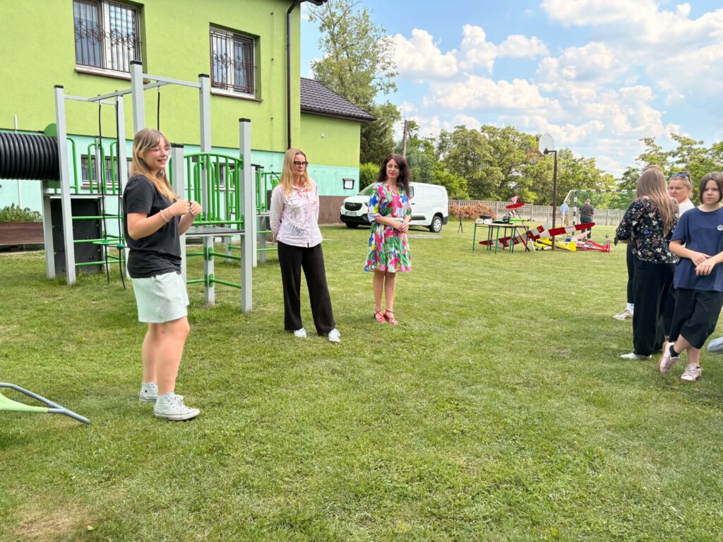 Grupa osób stoi na trawie obok placu zabaw i stołów z modelami samolotów na tle zielonego budynku i nieba z chmurami.