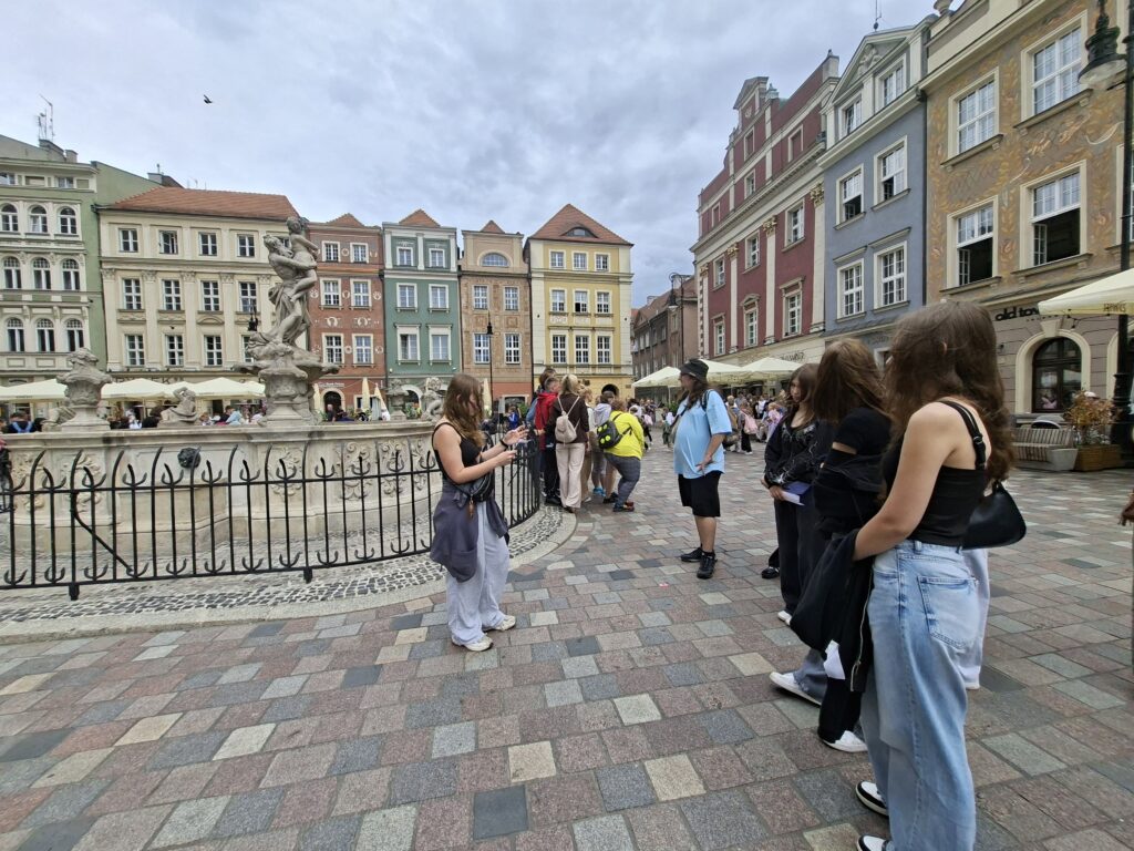 Tłum osób stoi w kolejce wzdłuż zabytkowej fontanny na kolorowym rynku otoczonym kamienicami o różnorodnych fasadach.