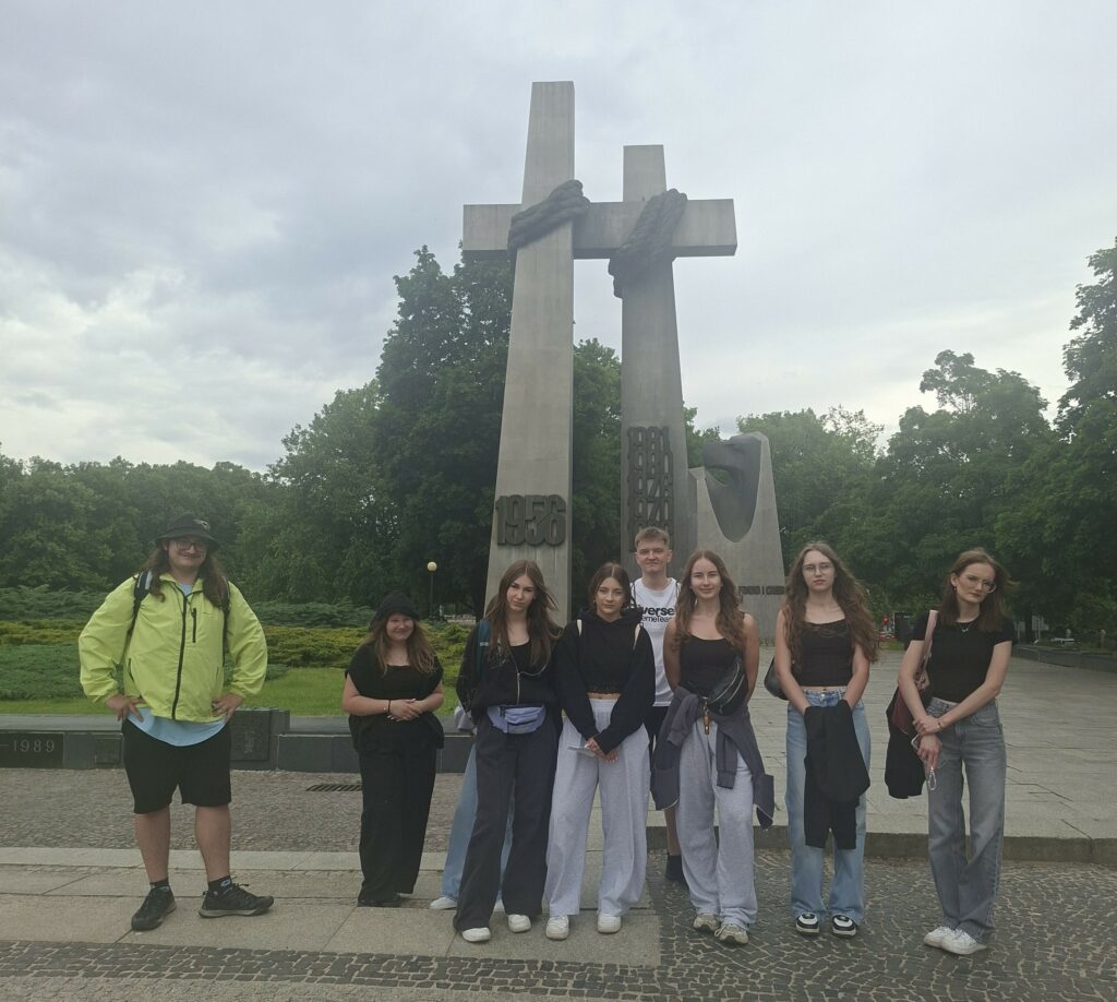 Grupa młodych ludzi stoi przed pomnikiem trzech krzyży, upamiętniającym ważne daty historyczne, na tle zielonych drzew i pochmurnego nieba.