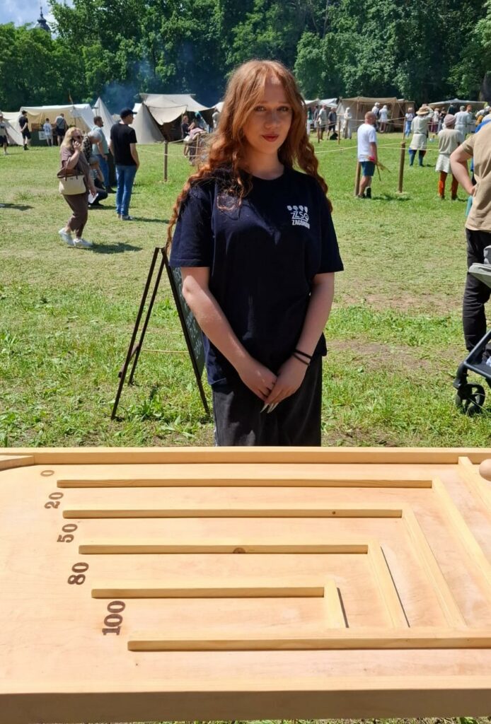 Młoda kobieta stoi za dużą drewnianą planszą z oznaczeniami punktacji, na tle osób i namiotów na otwartej przestrzeni.