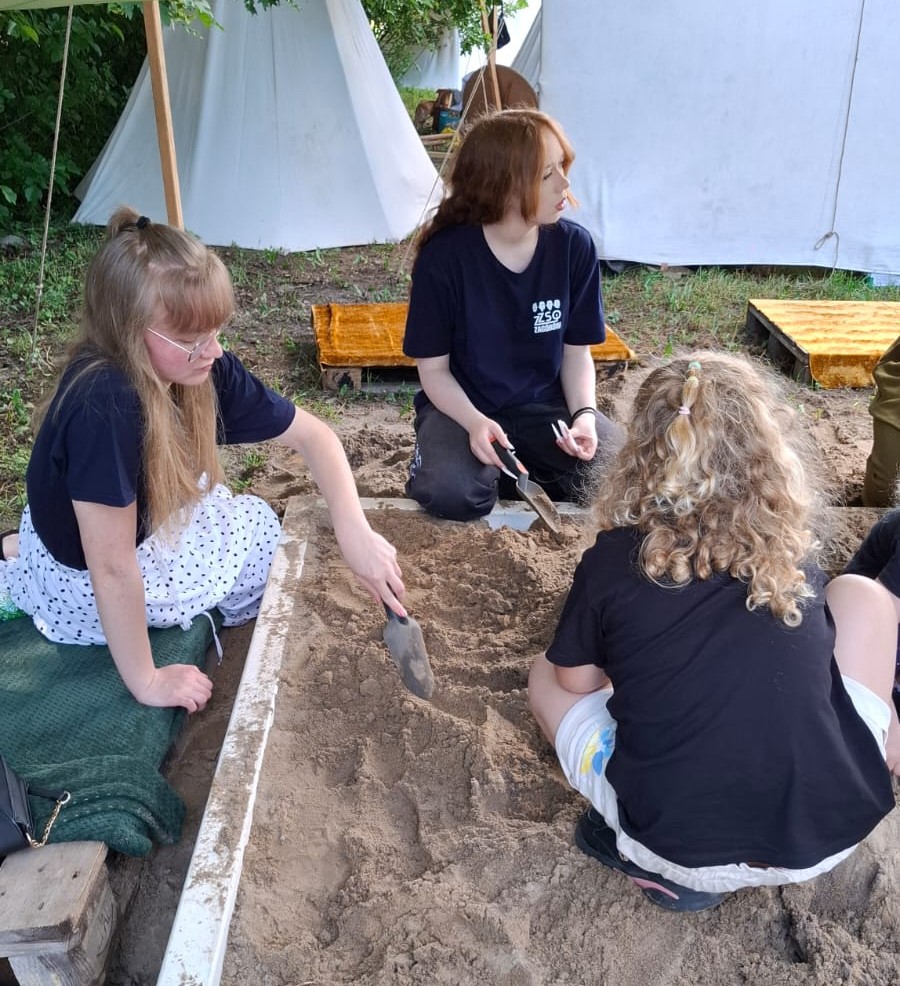 Grupa młodych osób kopie i przeszukuje piasek w archeologicznym wykopie na tle namiotów.