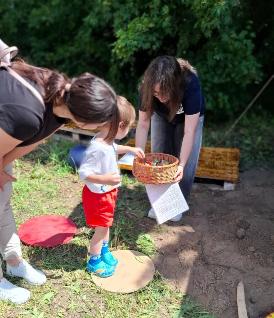 Dziecko stoi na okrągłej platformie na trawie, wybierając coś z koszyka trzymanego przez kobietę, obok stoi druga kobieta pochylona ku dziecku.