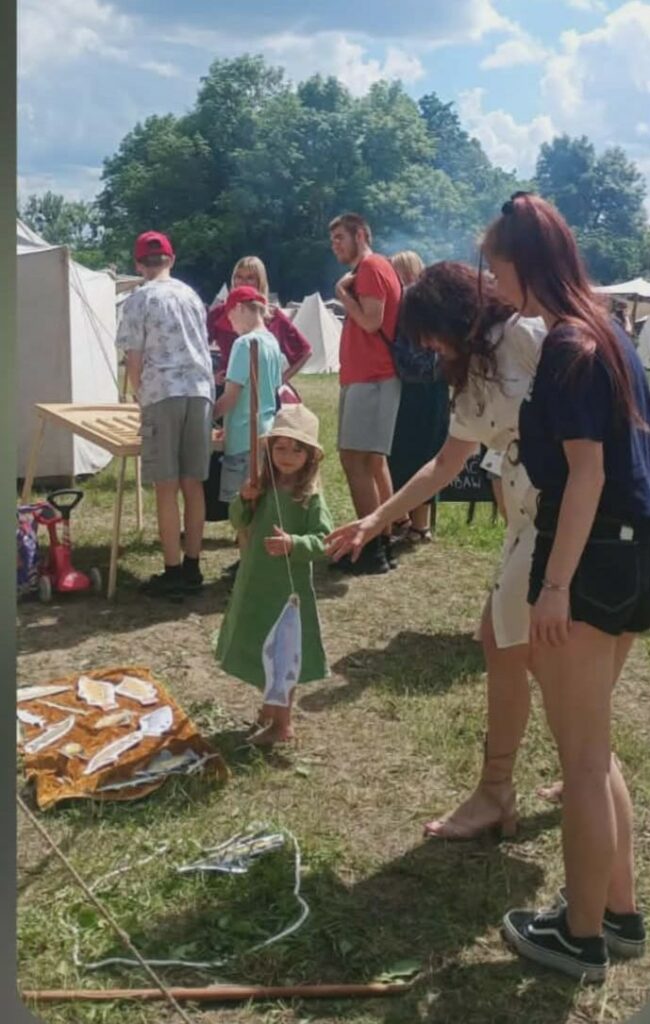 Dzieci i dorośli biorą udział w zabawie na świeżym powietrzu, łowiąc drewniane ryby za pomocą wędek z magnesem.
