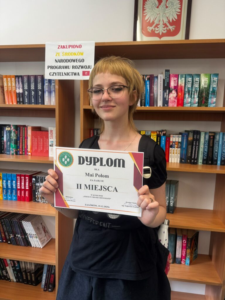 Dziewczyna z krótkimi włosami i okularami trzyma dyplom za zajęcie II miejsca w konkursie „Szkolny Mistrz Ortografii” w bibliotece szkolnej.