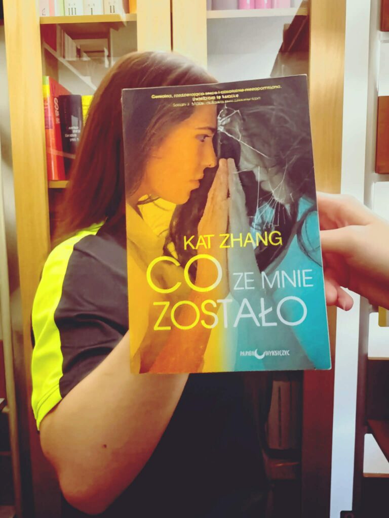 Książka „Co ze mnie zostało” Kat Zhang z ilustracją dwóch kobiet patrzących na siebie, jedna po stronie kolorowej, druga po stronie czarno-białej, trzymająca dłonie w geście dotyku.