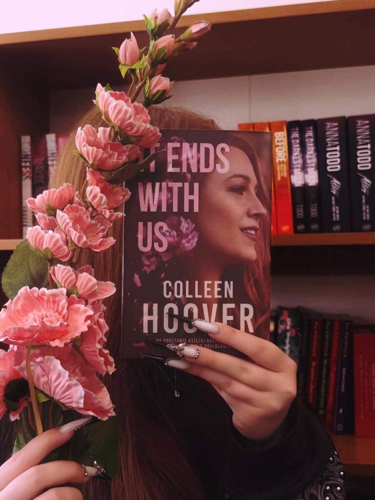 Ręce z długimi paznokciami trzymają książkę Colleen Hoover „It Ends With Us” oraz gałązkę z różowymi kwiatami.