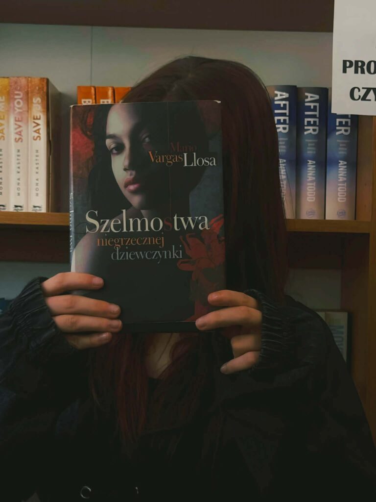 Książka "Szelmostwa niegrzecznej dziewczynki" Mario Vargas Llosa zasłania twarz osoby stojącej przy regale z innymi książkami.