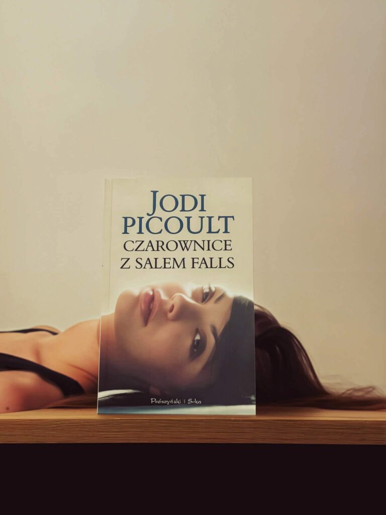 Książka „Czarownice z Salem Falls” Jodi Picoult na drewnianym stole, z twarzą kobiety w tle tworzącą iluzję połączenia z okładką.