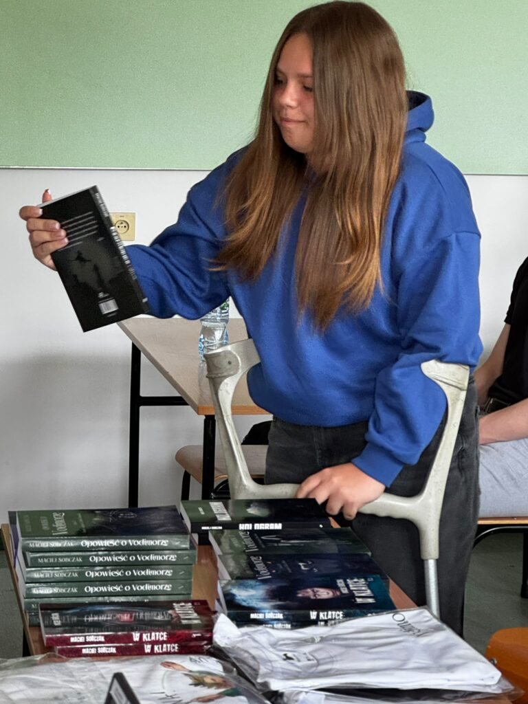 Dziewczyna w niebieskiej bluzie korzysta z kul łokciowych, wybierając książkę ze stołu pełnego różnych tytułów.