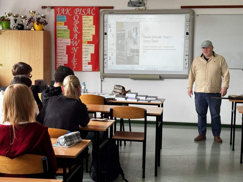 Mężczyzna prowadzi prezentację o pisarzu Macieju Sobczaku przed słuchaczami w klasie. Obok widoczna plansza edukacyjna z zasadami poprawnej pisowni.