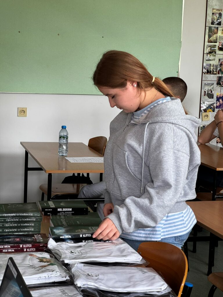 Młoda dziewczyna w szarej bluzie stoi przy stole z poukładanymi książkami i białymi koszulkami w folii.