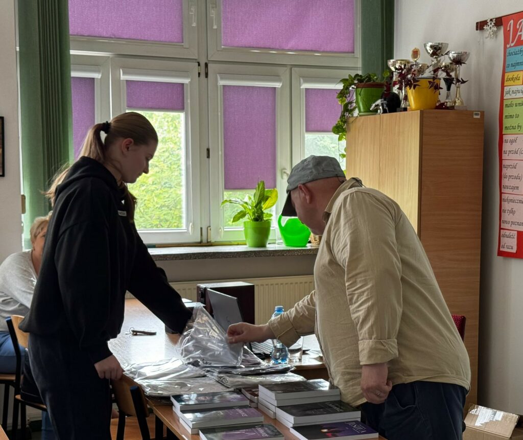 Dwie osoby oglądają książki i pakiety na stole w dobrze oświetlonym pomieszczeniu z fioletowymi roletami.