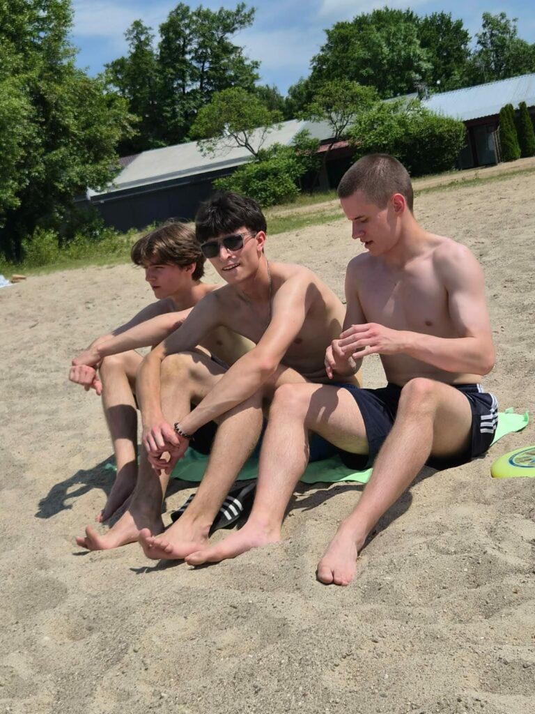 Trzech młodych mężczyzn w kąpielówkach odpoczywa na piasku w słoneczny dzień, w tle drzewa i budynki.
