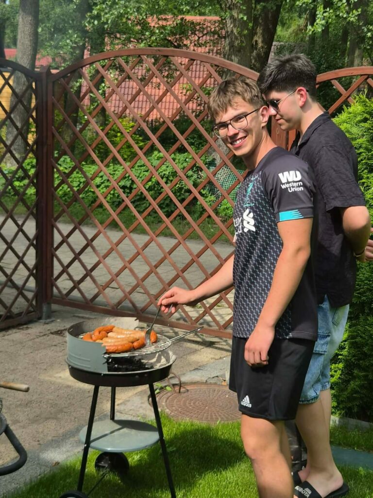 Dwóch młodych mężczyzn przy grillu, jeden z nich obraca kiełbaski na ruszcie.