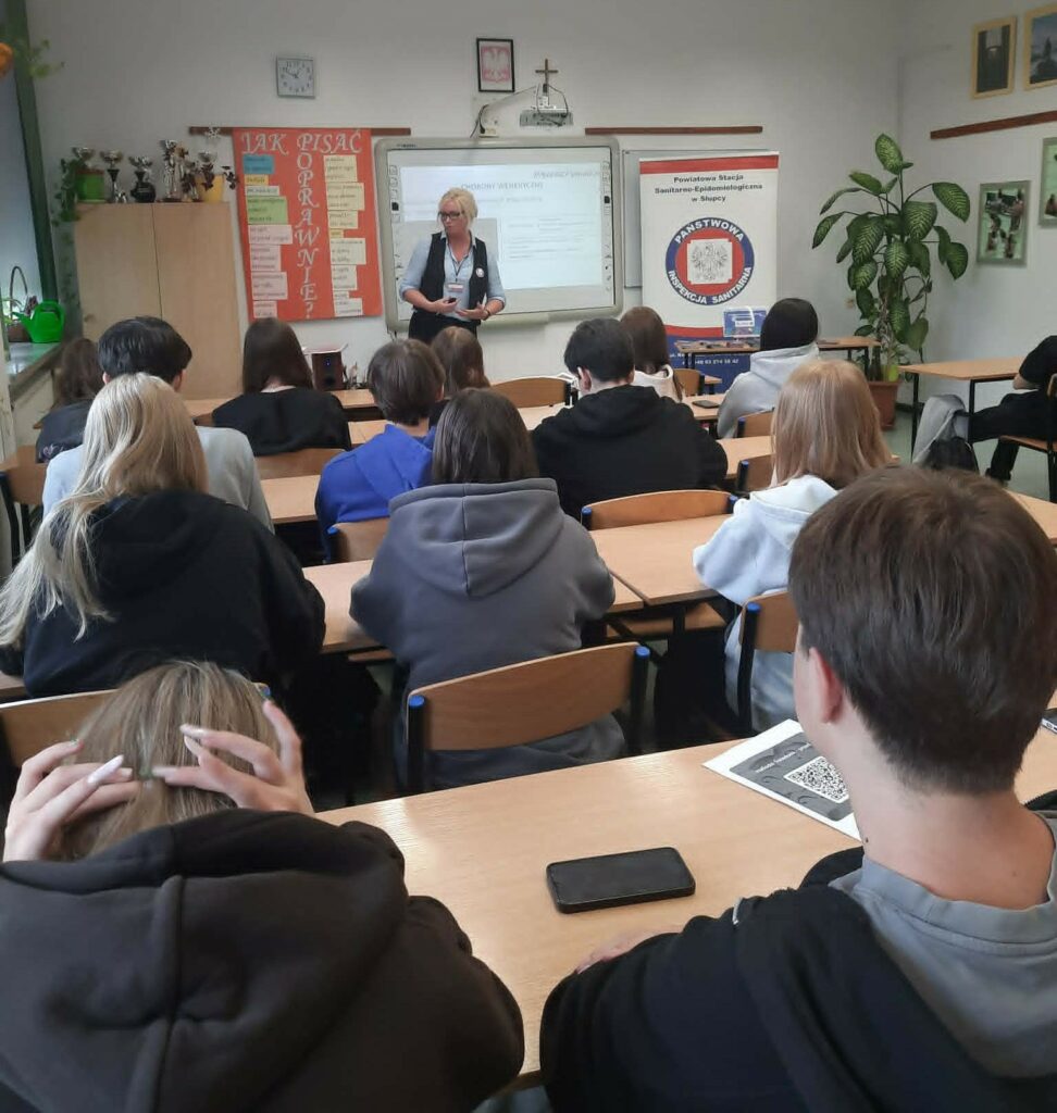 Grupa uczniów uczestniczy w lekcji prowadzanej przez kobietę na temat chorób wenerycznych w klasie wyposażonej w tablicę interaktywną i materiały edukacyjne.