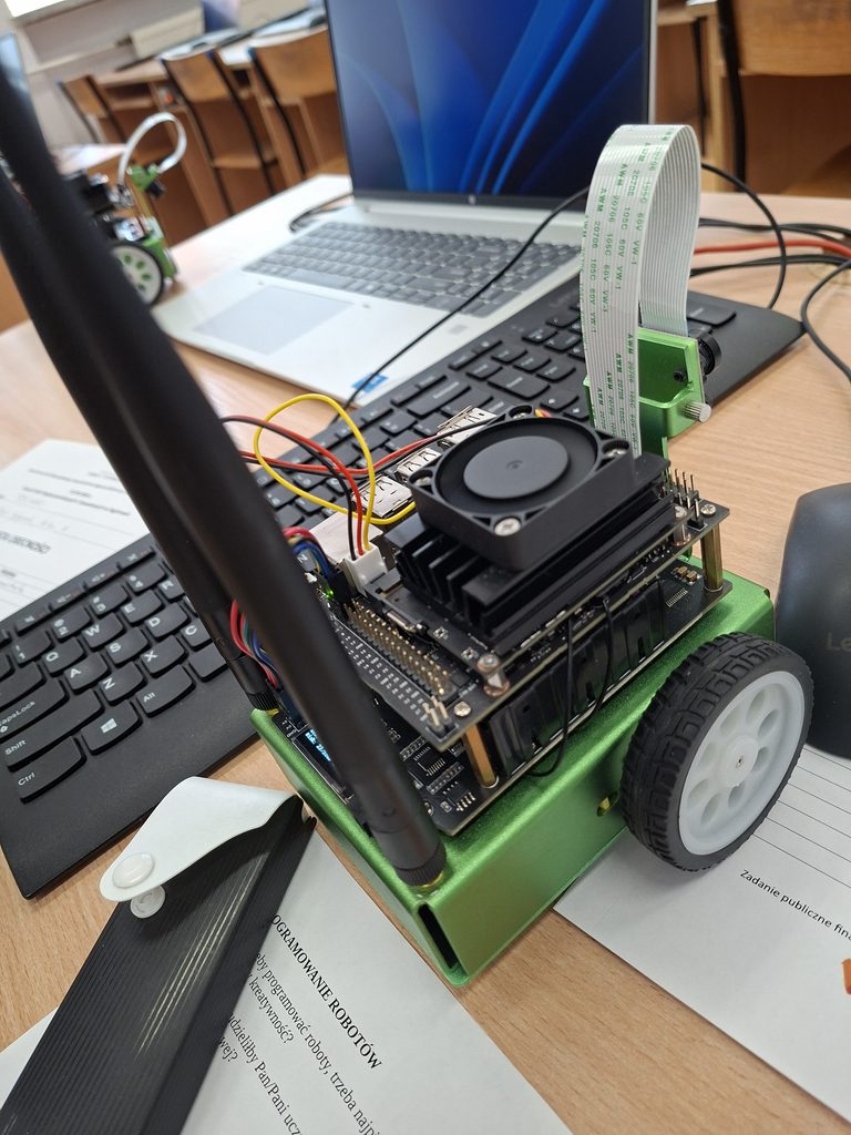 Komputer z podłączonym modułem elektronicznego robota z dwoma kołami i kamerą, ustawiony na biurku obok laptopa i dokumentów.